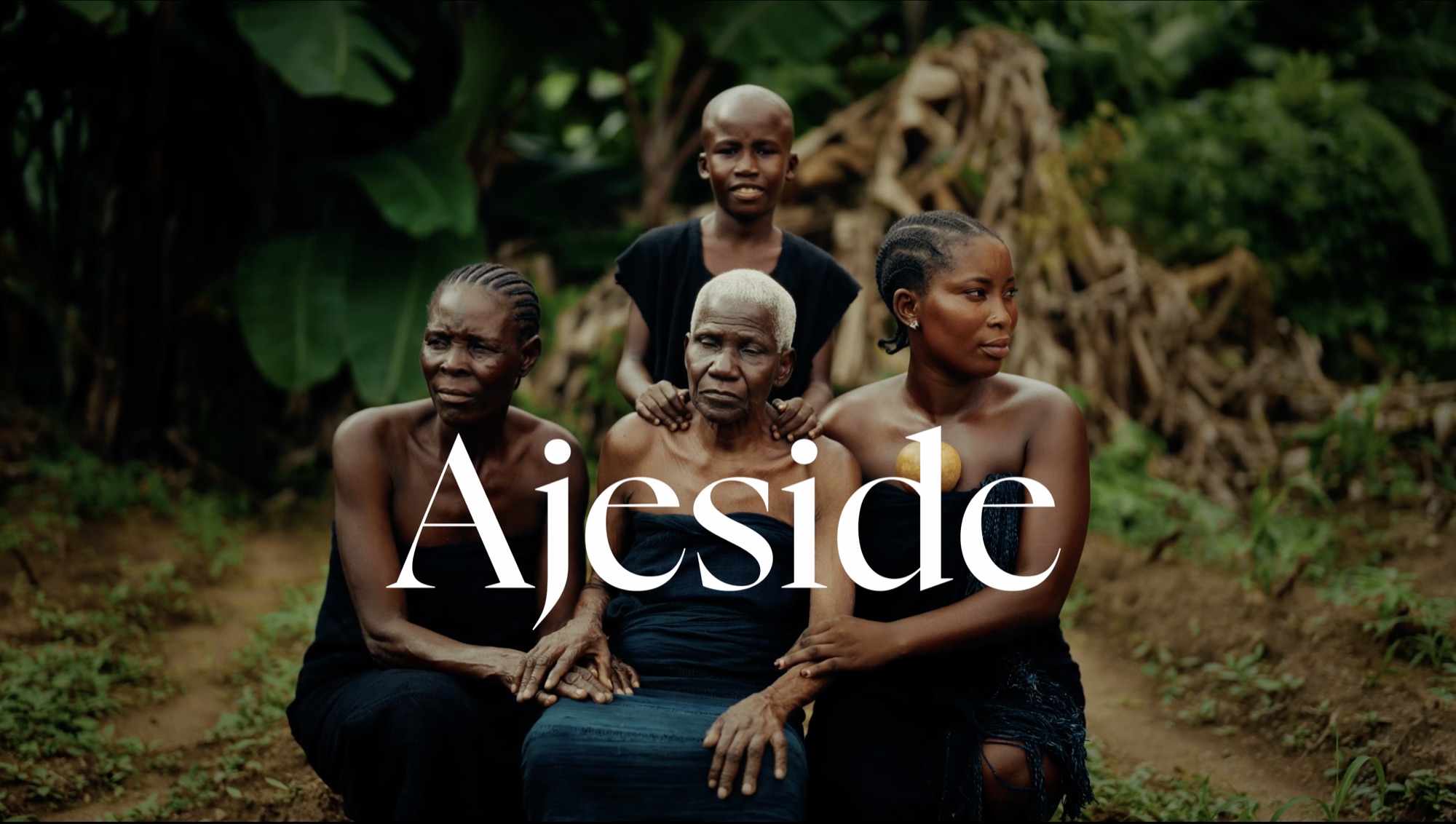 Four generations of a Gbagyi family seated together in dark indigo cloth against dense green foliage