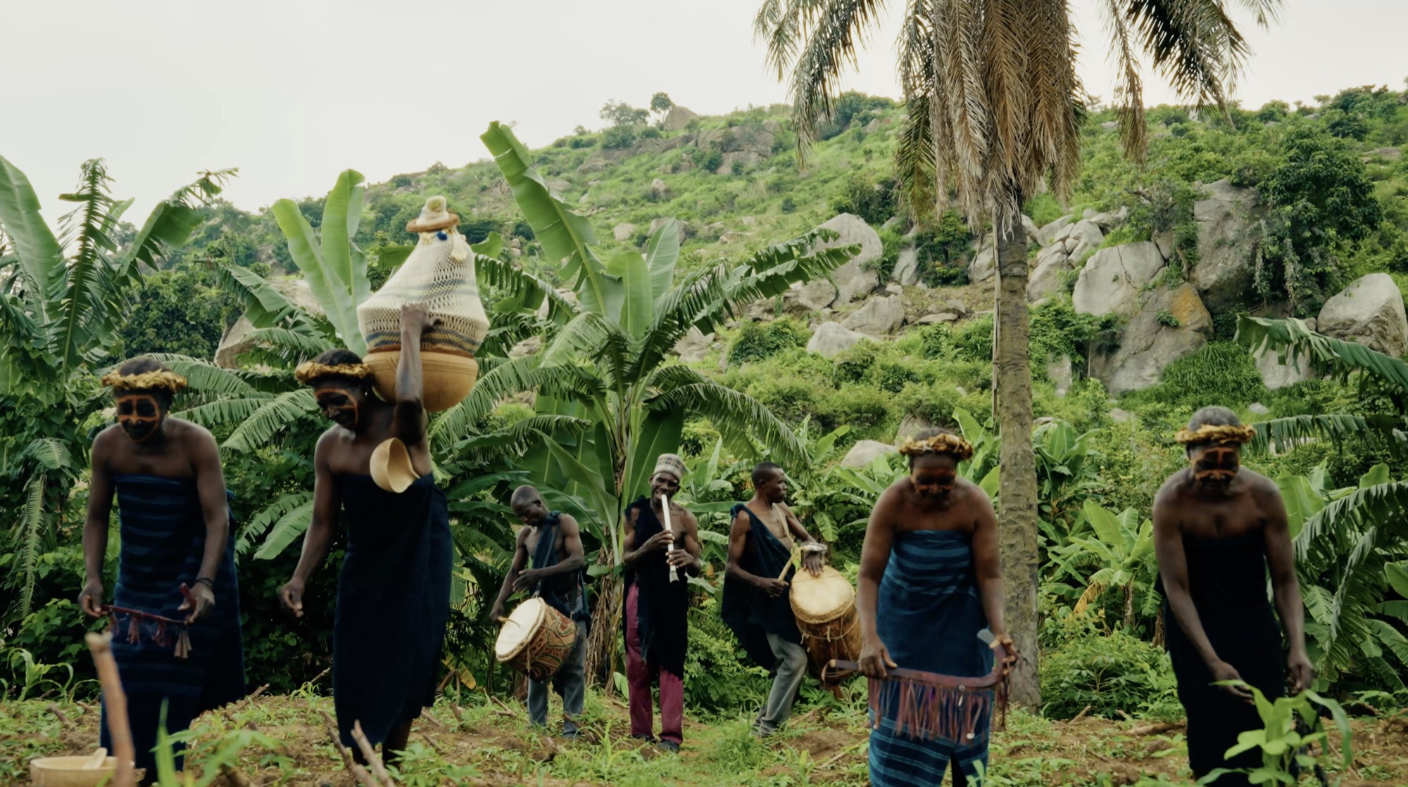 Women carrying woven vessels lead a procession of drummers and flute players through banana palms below a rock outcrop