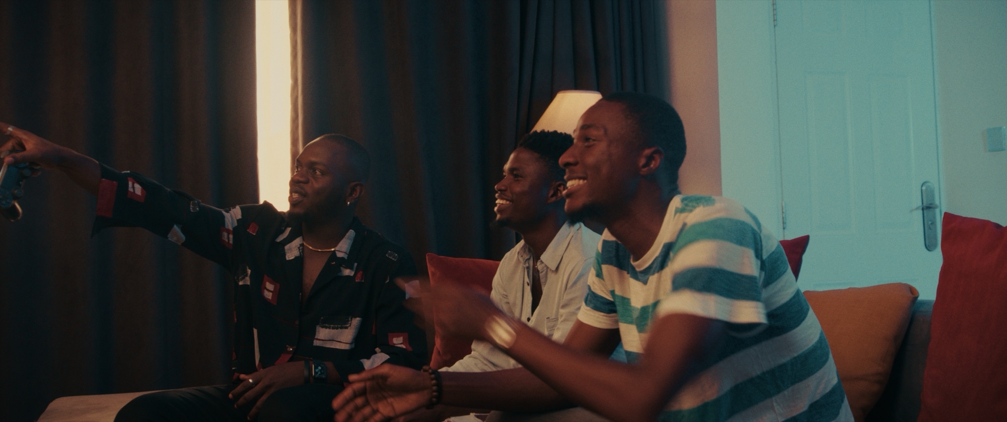 Three friends sitting along a sofa in warm lamplight, laughing while playing a video game
