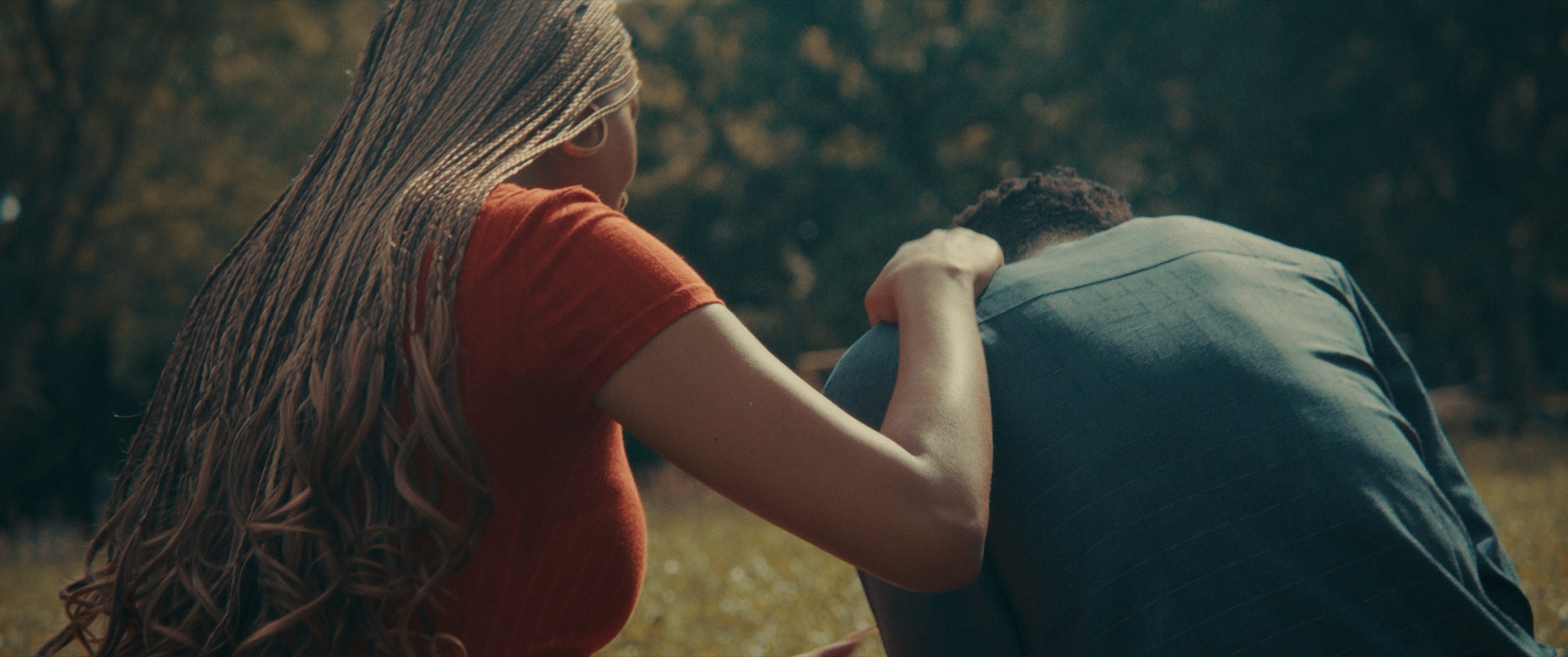 Seen from behind, a woman in a red top rests her hand on the shoulder of a man bent forward beside her