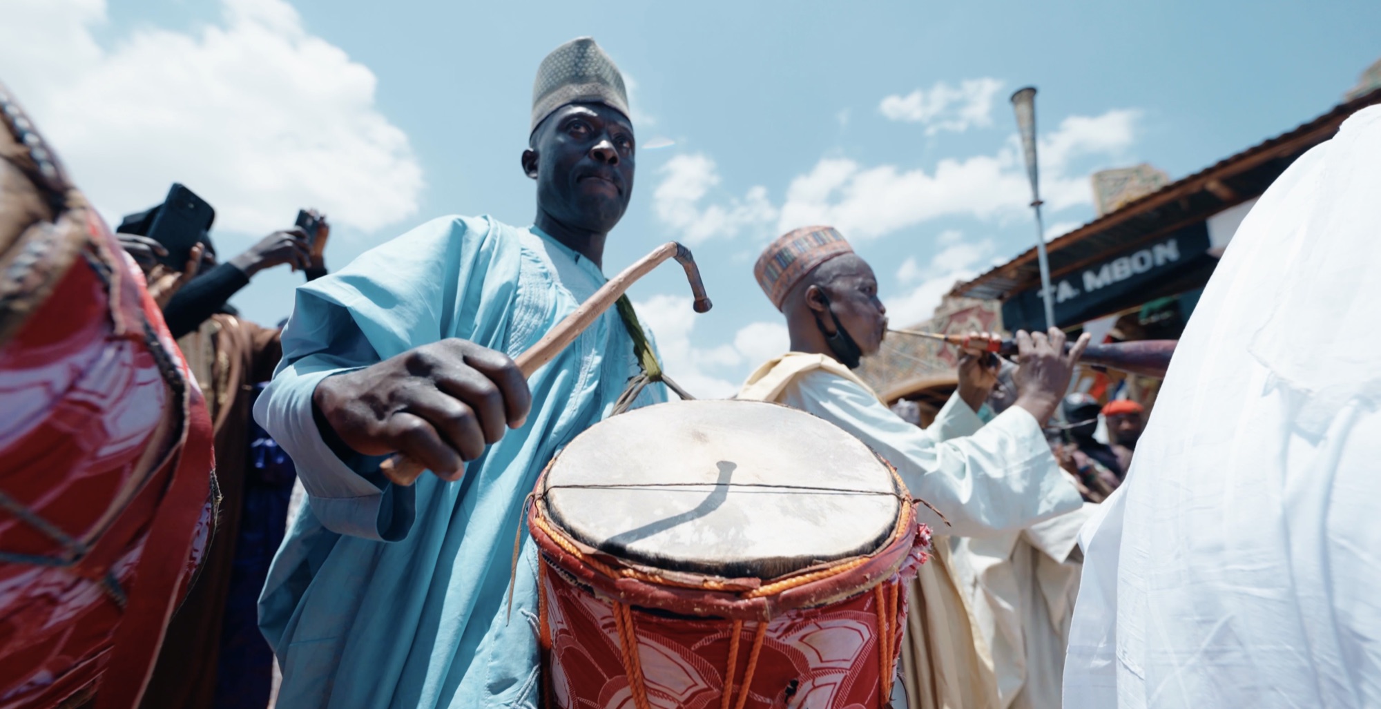 Drummers in pale blue robes and embroidered caps playing in a crowded street beneath a bright sky