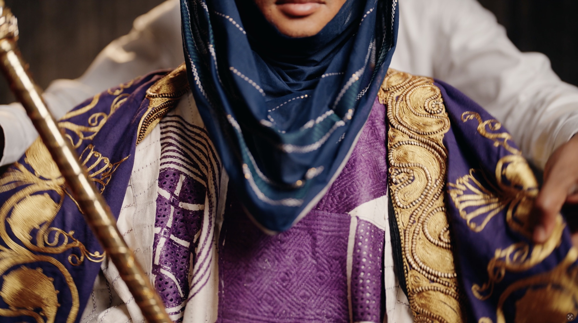Close view of ceremonial dress: gold embroidery on purple and white cloth, with a deep blue head covering