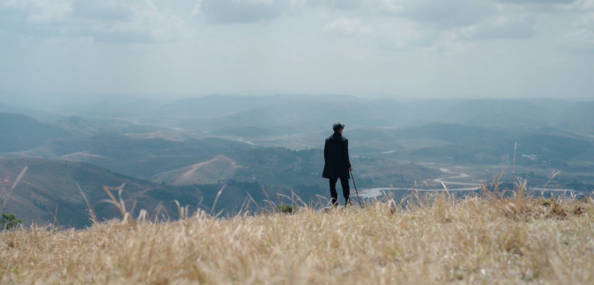 A lone figure in a dark robe stands in tall dry grass on a hilltop, looking out over the valleys of Mambilla