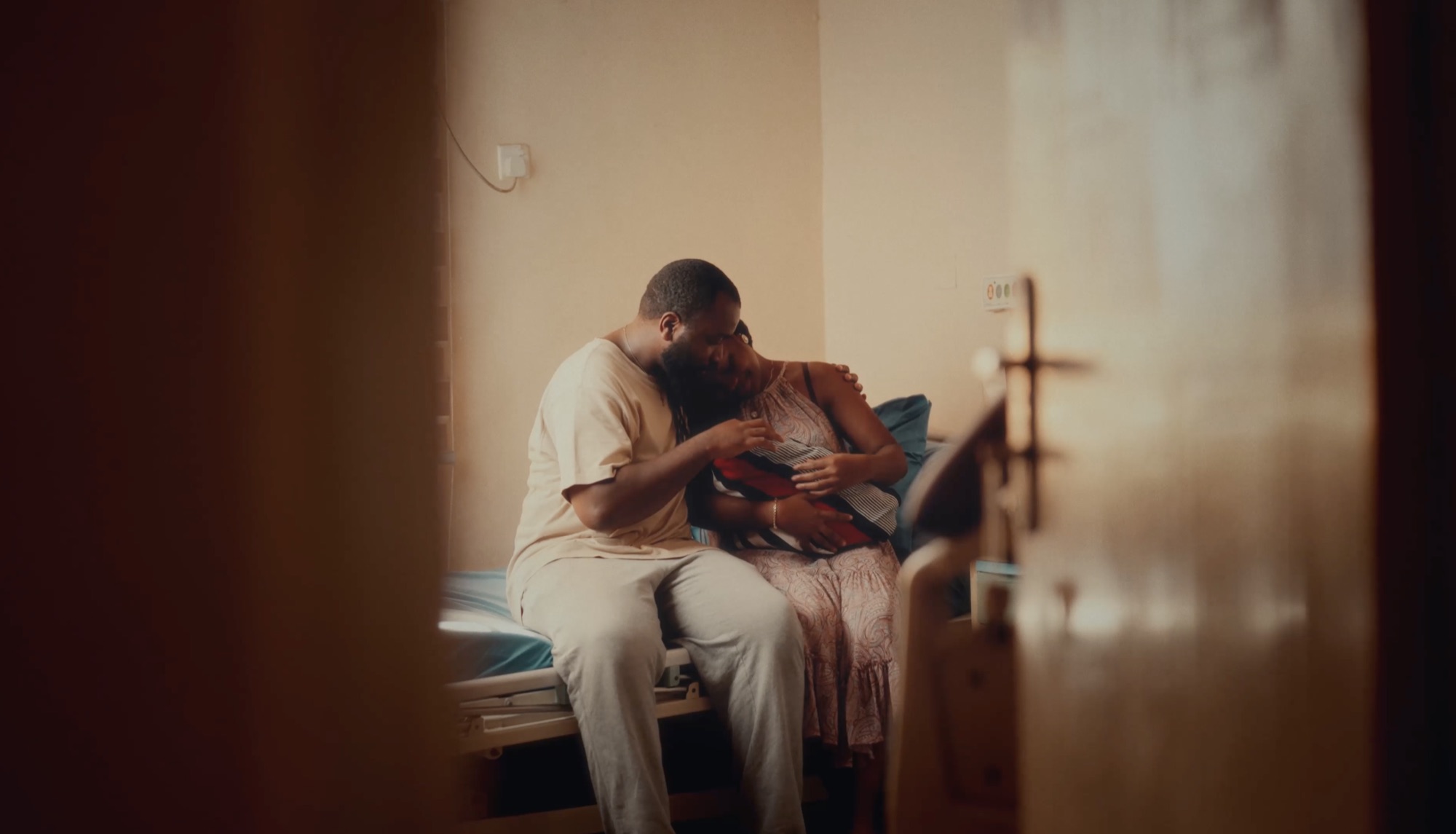 A man sits on a hospital bed with his arm around a woman, the two leaning into each other in warm afternoon light