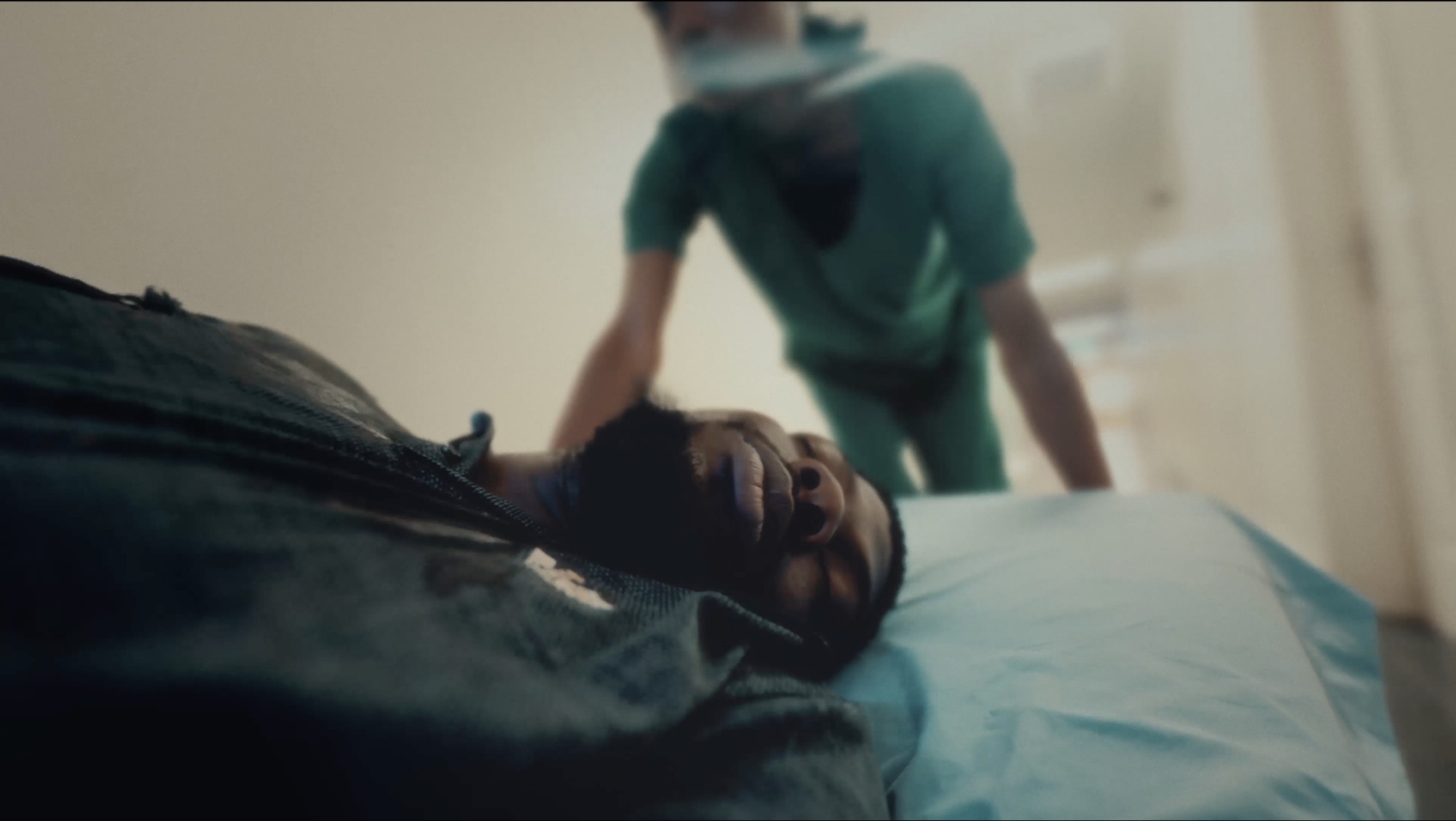 A patient lies on a hospital bed as a masked clinician in green scrubs leans over them